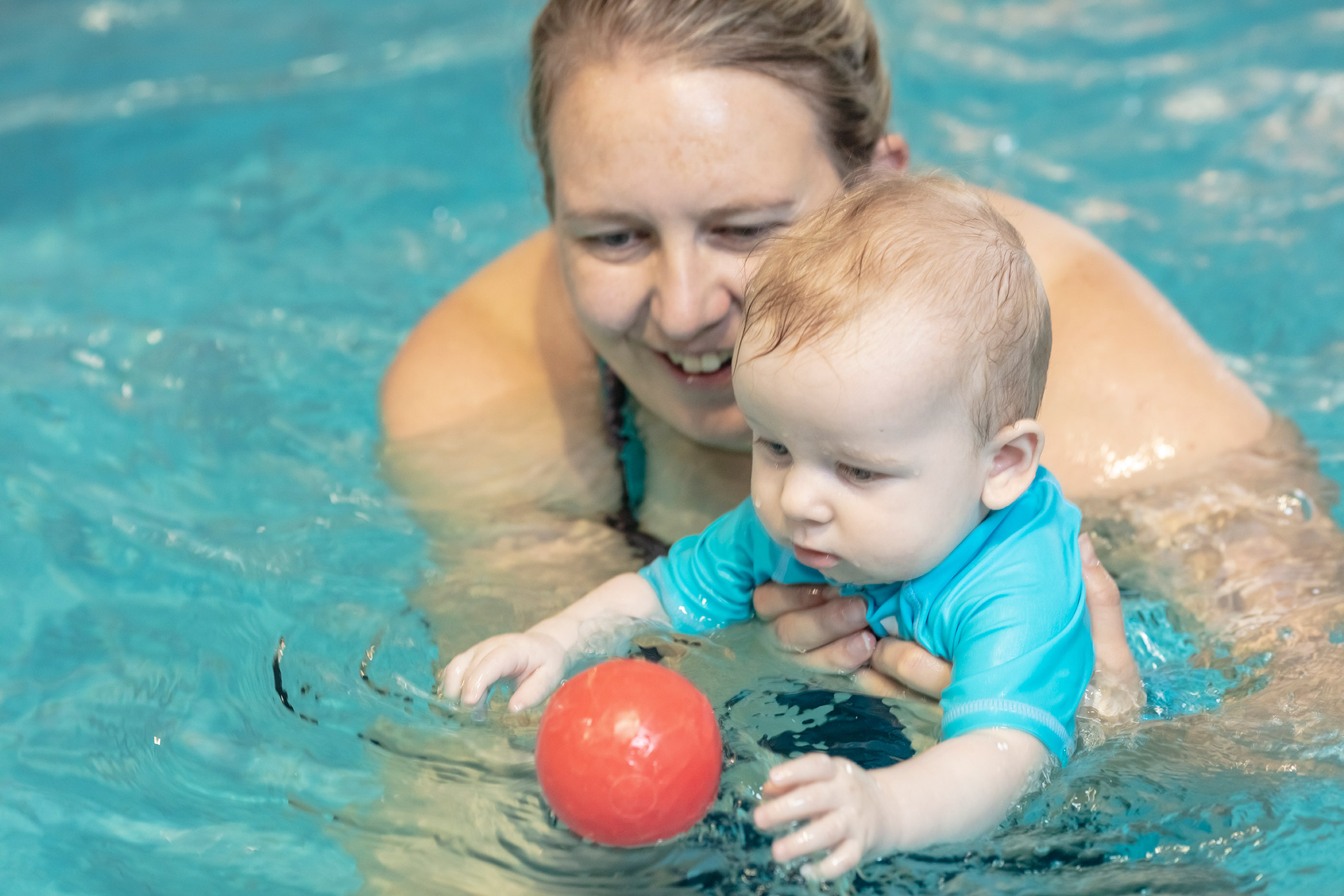 A toddler reaching out for a red ball while being held by a smiling woman in a Christchurch Recreation and Sport pool. 