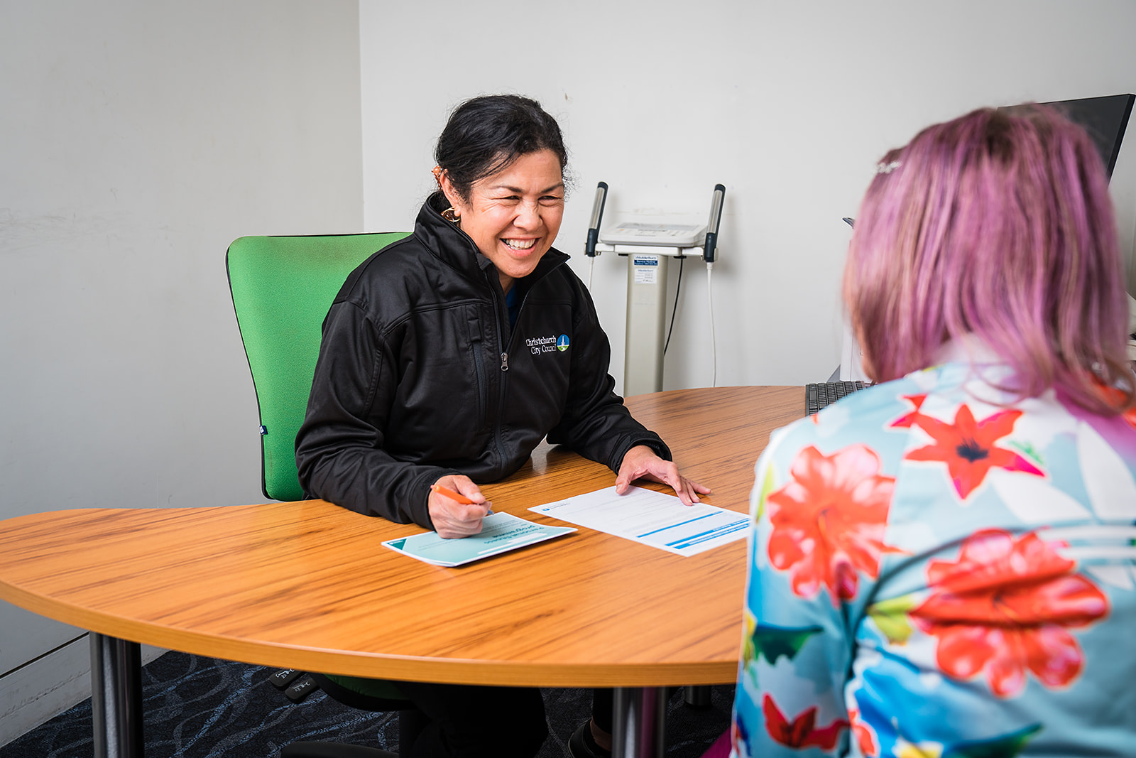 Two smiling women seated at a desk. One woman, a personal trainer is seated across from a woman wearing floral and pink hair. 