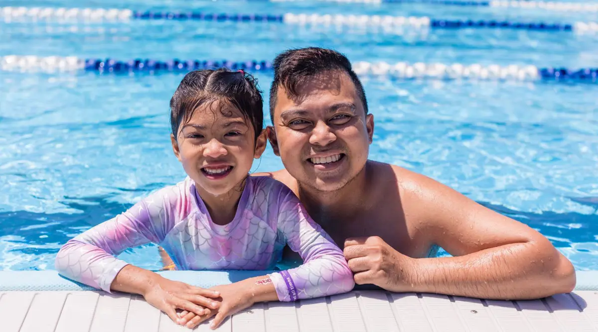 A smiling little girl wearing pink swimmers and a smiling man swimming at the Halswell Summer pool.