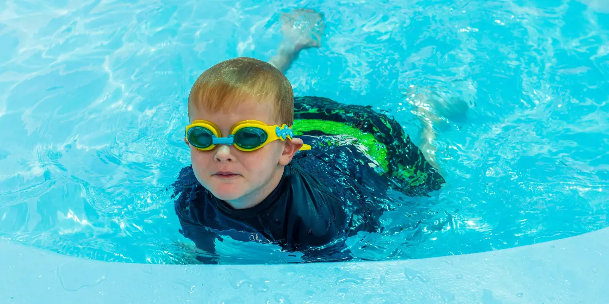 A little boy in yellow goggles and blue swimmers in the Waltham Summer toddler pool.
