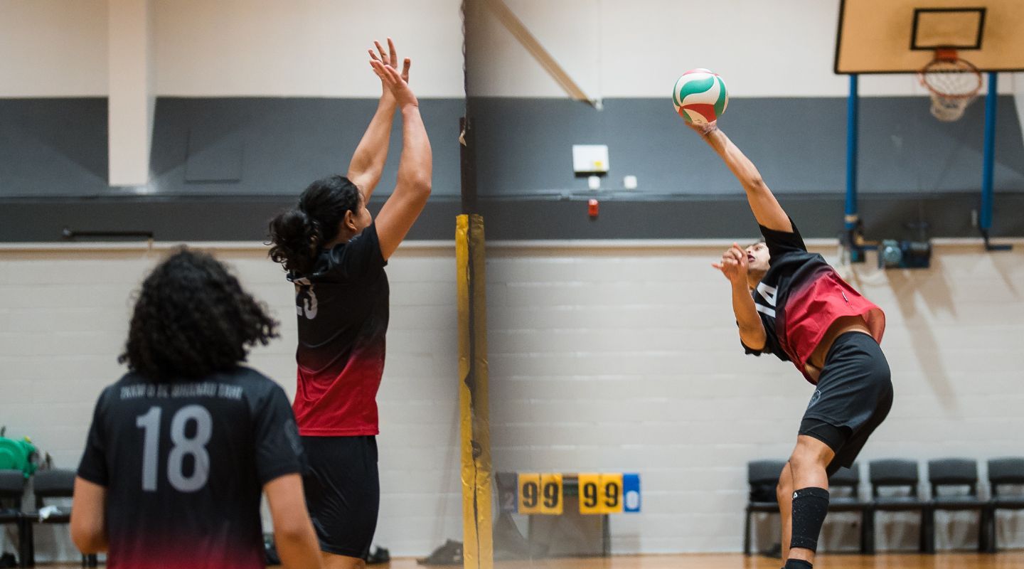 Volleyball players in red uniforms jump up on either side of the net as one spikes, and one blocks the ball at Christchurch Recreation and Sport centre.  