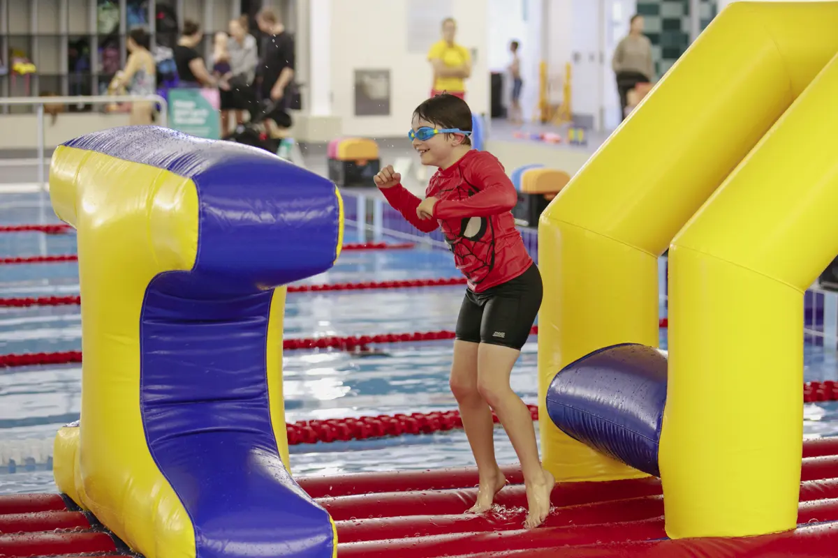 A smiling little boy jumping along one of the inflatable obstacle courses at Mataiki Hornby pool.