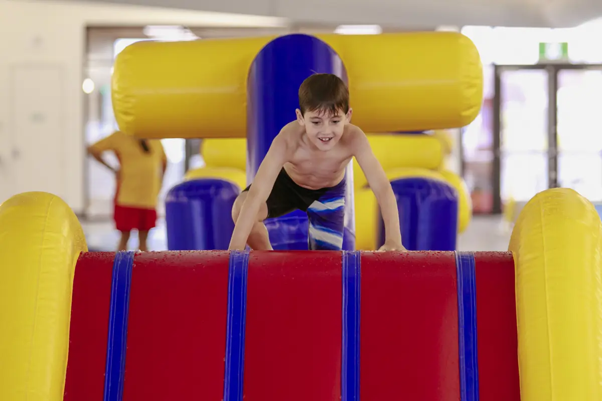 A smiling little boy climbing up to the top of the inflatable obstacle course at Mataiki Hornby pool.
