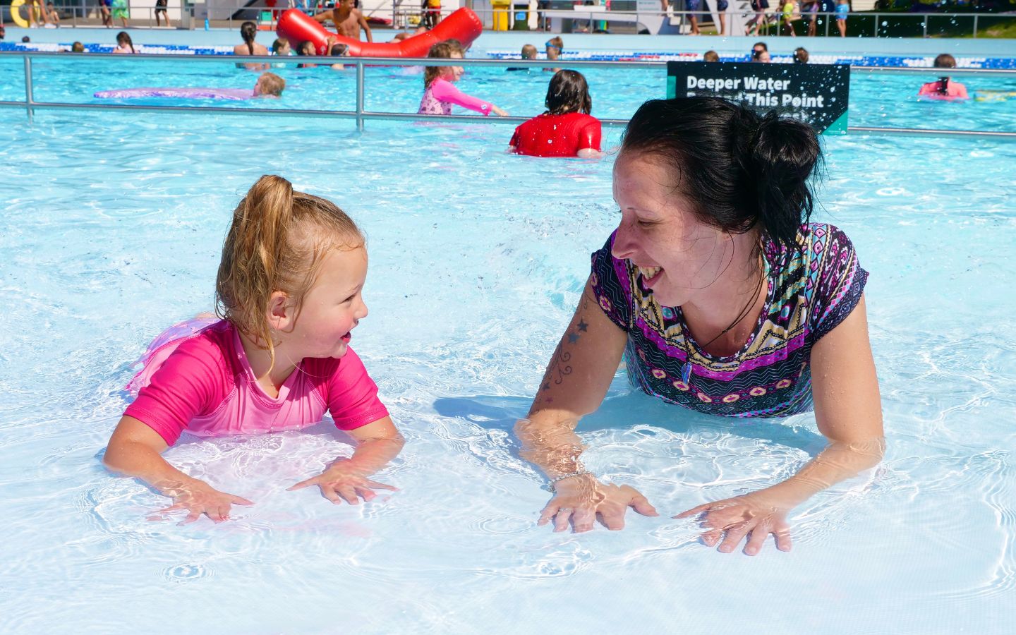 A child in pink and a smiling woman lay on their stomachs in the toddler pool at a Christchurch summer pool. 