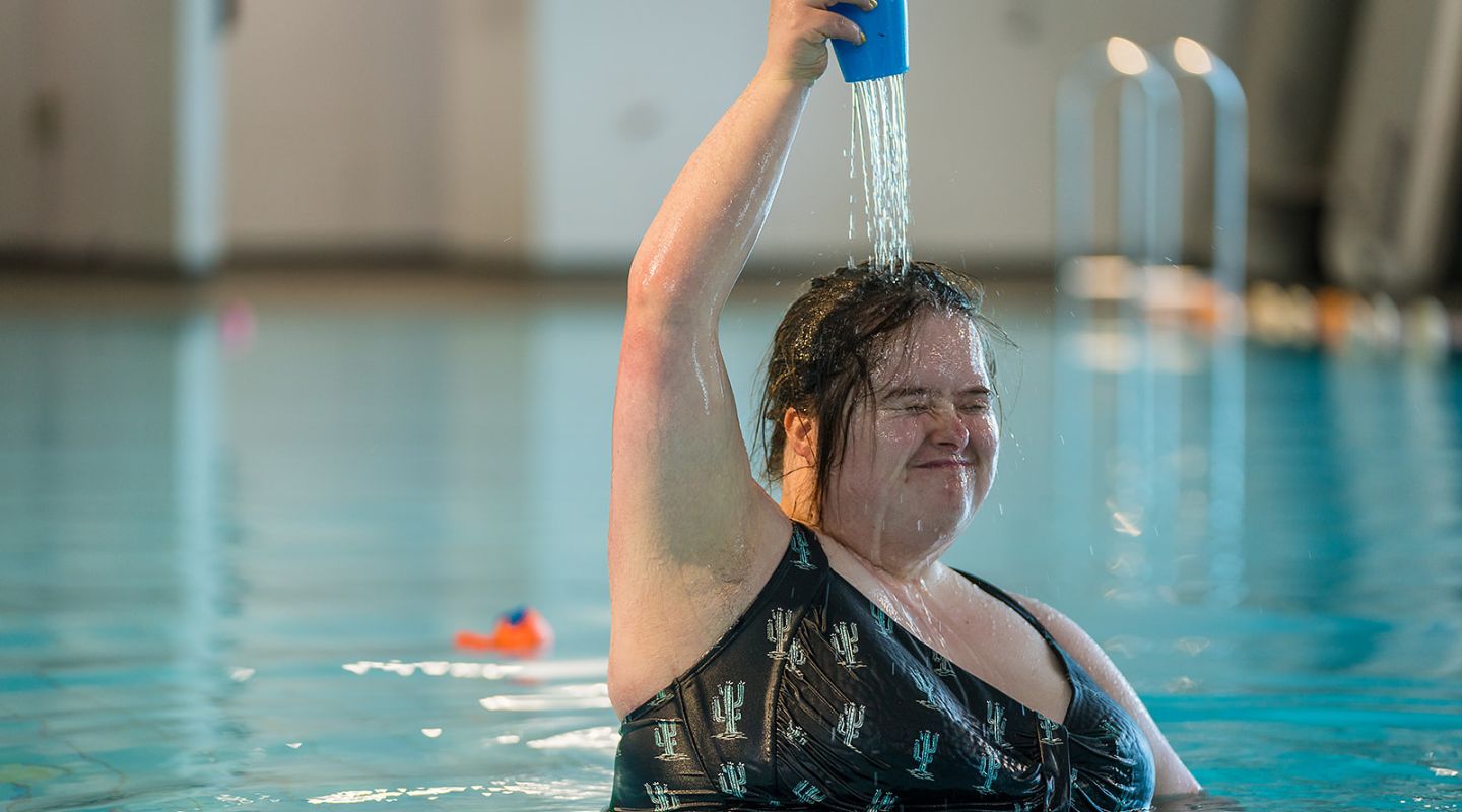 A smiling woman playing with a blue cup pouring water on her head during a Sensory Swimming class at Christchurch Recreation and Sport pool.