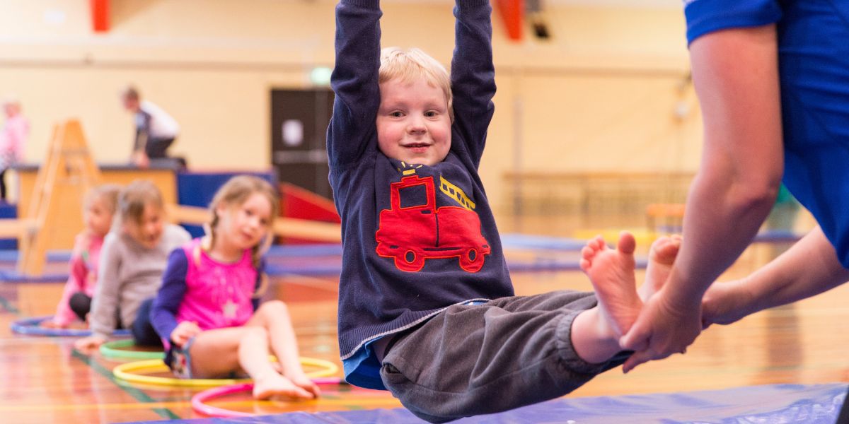 A smiling child holding onto hanging rings during a preschool gym class at Pioneer Recreation and Sport centre. 