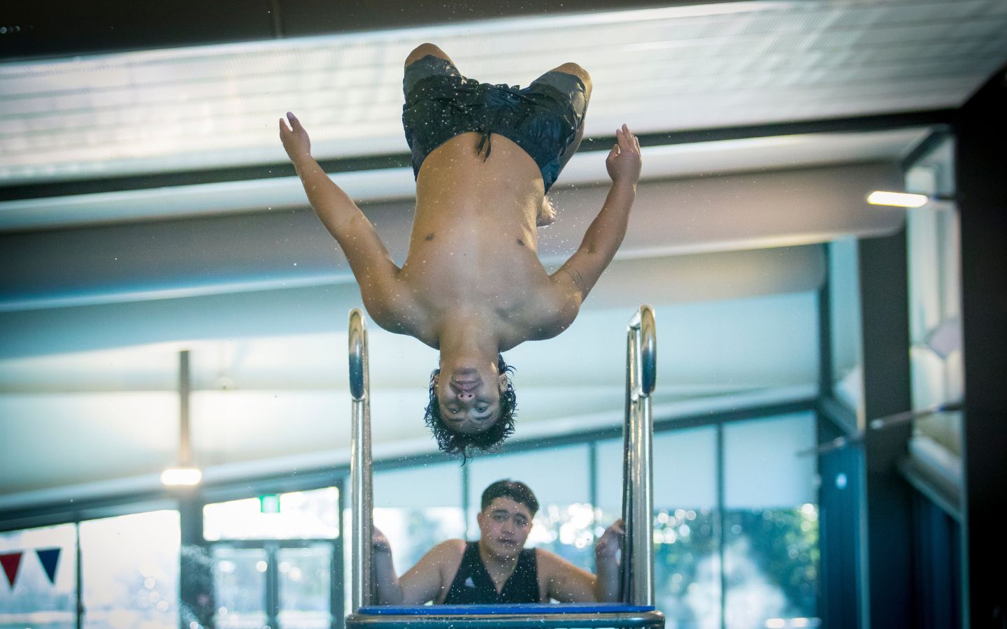 A teenage boy doing a backflip off a diving board at Christchurch Recreation and Sport pool. 