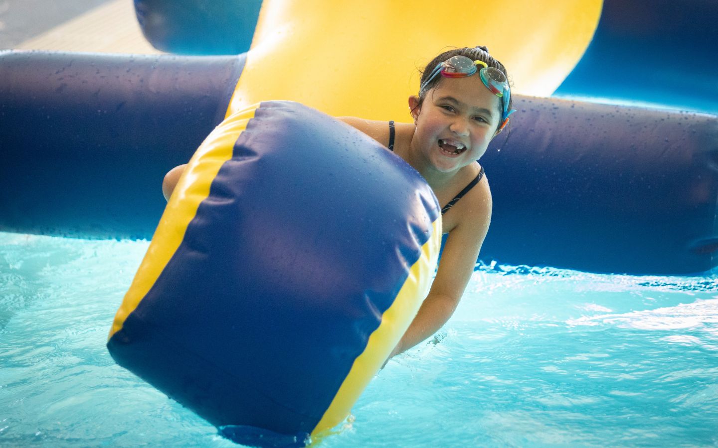 A smiling girl playing on a yellow and blue inflatable at the Christchurch Recreation and Sport pool. 