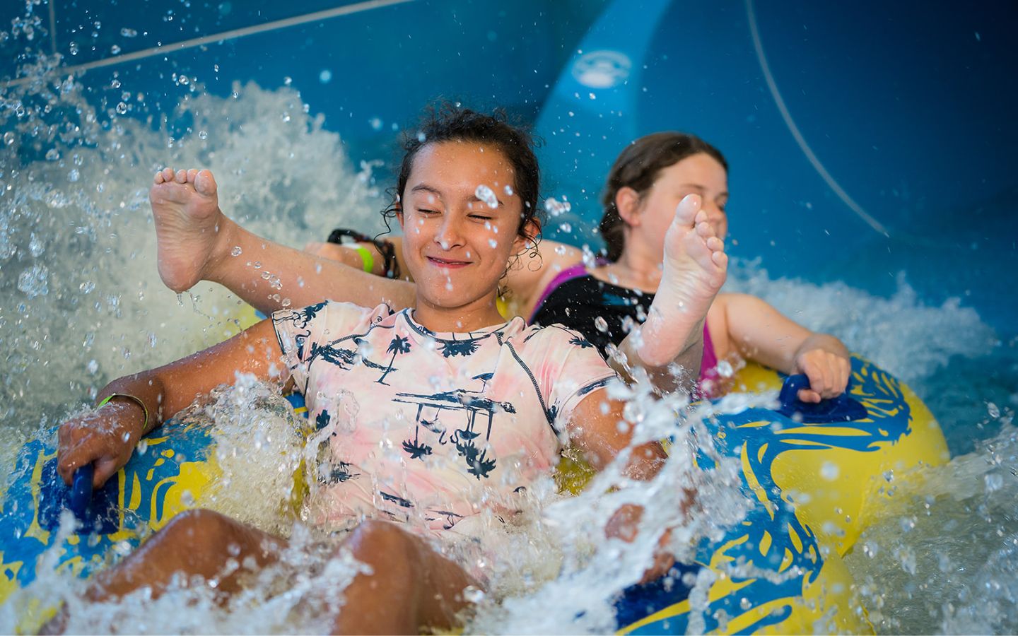 Two smiling girls sliding down the blue hydroslide at QEll Recreation and Sport Centre in a yellow tandem raft. 