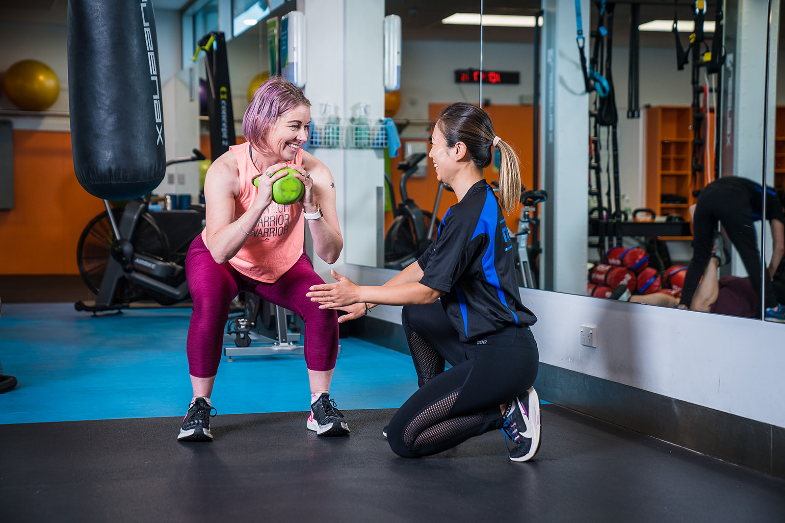 A female personal trainer assisting a smiling woman wearing pink while she does a squat at Pioneer Rec Centre. 